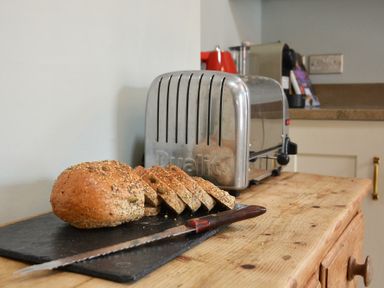 A sliced loaf of seeded bread on a cutting board with a serrated knife and a metal toaster on a wooden kitchen counter at Walton House Southwold