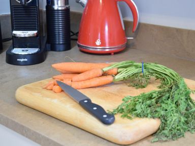 A kitchen countertop with a wooden cutting board holding a bunch of carrots some sliced carrots and a knife with a black handle next to a red electric kettle and a black coffee machine at Walton House Southwold