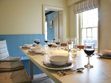 A dining table set with bowls glasses of red wine and a water jug in a room with a window and blue bench seating at Walton House Southwold