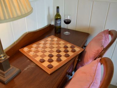 A wooden table with a checkerboard and pieces two wooden chairs with pink cushions a bottle and glass of red wine with a lamp on the side at Walton House Southwold