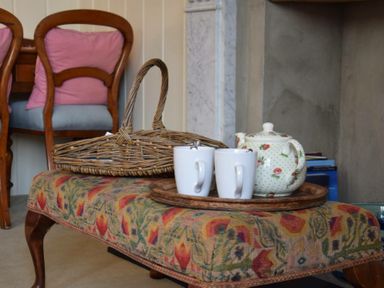 A wicker basket and a wooden tray with a floral teapot and two white mugs on a patterned ottoman in a room with wooden chairs at Walton House Southwold