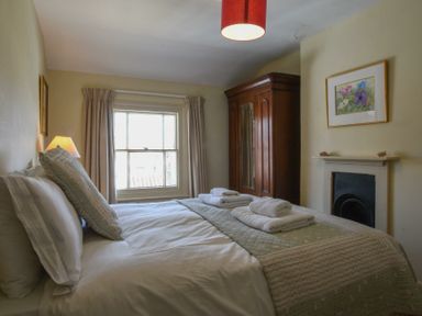 A bedroom with a bed, folded towels, a wooden wardrobe, a window with curtains, a fireplace, and a framed picture on the wall at Walton House, Southwold