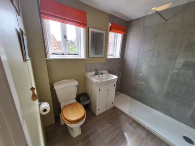 A bathroom with a toilet wooden seat a sink under two windows with orange blinds and a shower area at Walton House Southwold