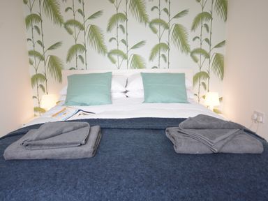 A bed with folded towels on a blanket and pillows against a wall with palm leaf wallpaper at Sweet Pea Southwold