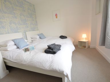 A bedroom with two single beds, white bedding, blue pillows, folded dark towels, a small white bedside table with a lamp and books, and blue wallpaper at Sweet Pea, Southwold