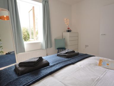 A bedroom with a bed folded towels on a blanket a window with curtains a chest of drawers and a chair with a cushion