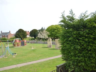A playground with climbing equipment and a slide near a path with trees and houses in the background at Sweet Pea Southwold