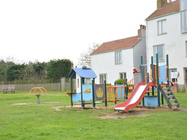 A playground with slides and climbing equipment in a grassy area near white houses with tiled roofs in Southwold