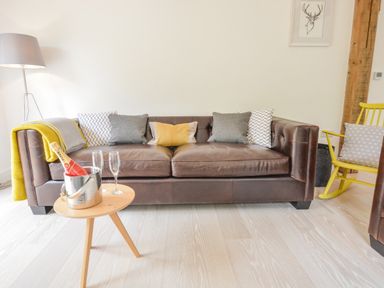 A brown leather sofa with cushions and a yellow throw next to a wooden side table with a bottle in an ice bucket and two glasses at Spexhall Barns in Halesworth