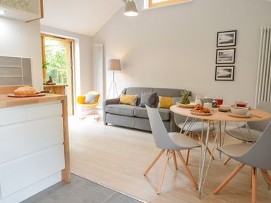 A dining area with a round table set for breakfast and a gray sofa with cushions in a living room at Spexhall Barns in Halesworth