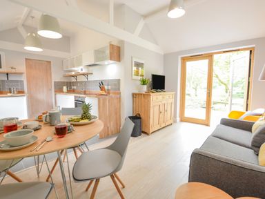 A kitchen and living area with wooden dining table set for breakfast a grey sofa and wooden cabinet with television at Spexhall Barns in Halesworth