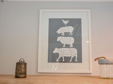 A framed diagram of meat cuts from a chicken pig sheep and cow on a wall above a wooden shelf with a metal container and a glass jar with a rope handle at Spexhall Barns in Halesworth