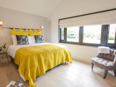 A bedroom with a bed covered in a yellow blanket and pillows beside a window with a gray chair near the window at Spexhall Barns in Halesworth