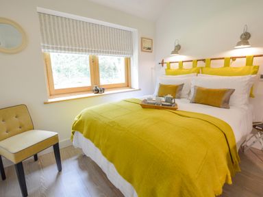 A bedroom with a bed covered with a yellow blanket yellow cushions a chair and a window at Spexhall Barns in Halesworth