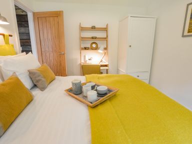 A bedroom with a bed, yellow pillows, a yellow blanket, a wooden door, a white wardrobe, and a small desk with a lamp and chair at Spexhall Barns in Halesworth