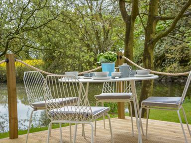 A round white table with bowls cups and a teapot with four white metal chairs on a wooden deck by a river at Spexhall Barns in Halesworth