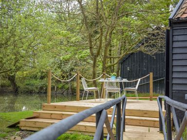 A wooden deck with a table and four chairs next to a pond and surrounded by trees at Spexhall Barns in Halesworth