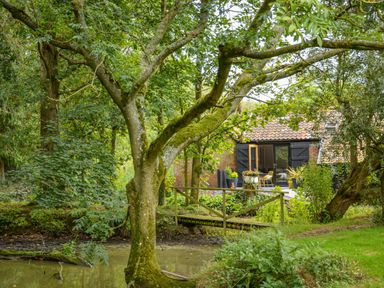 A garden with trees a small wooden bridge over a pond and a house with an open door and black shutters at Spexhall Barns in Halesworth
