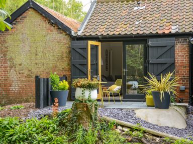 An outdoor patio with plants in pots and yellow chairs outside a brick building with black shutters at Spexhall Barns in Halesworth