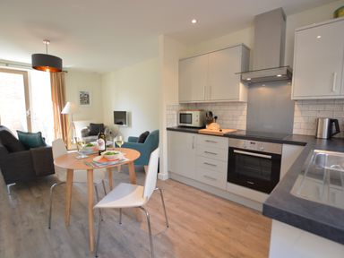 A kitchen and dining area with a round wooden table set for two and a living room with chairs and a small tv at Mycroft Southwold in Southwold