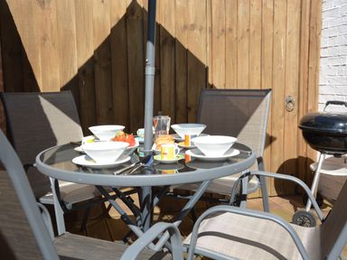 An outdoor patio dining area with a glass table set for four with bowls cups and strawberries beside a black barbecue grill at Mycroft Southwold