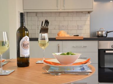 A kitchen table set with a bottle of white wine glasses a bowl of salad and cutlery with a kitchen counter with knives bread and a pot in the background at Mycroft Southwold in Southwold