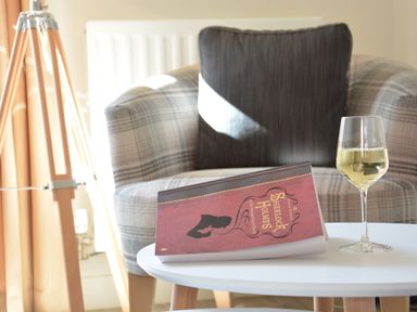 A plaid armchair with a dark cushion a white round table with a book and a glass of white wine beside a wooden tripod lamp at Mycroft Southwold in Southwold