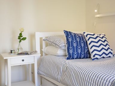 A white bed with striped blue and white bedding and patterned cushions next to a white bedside table with a vase with a flower at Mycroft Southwold in Southwold