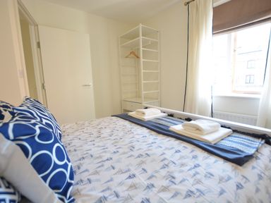 A bedroom with a bed with patterned bedding pillows a white shelving unit an open door and a window with curtains at Mycroft Southwold in Southwold