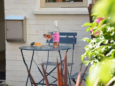 A small blue metal patio table and chair outside with a bottle of wine two glasses and a bowl of snacks at Mycroft Southwold