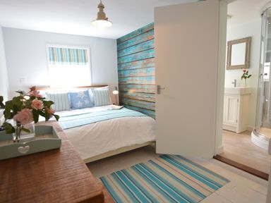 A bedroom with a bed with striped and starfish pillows a striped window blind a striped rug a wooden dresser with flowers and an open door leading to a bathroom at Alinka Aldeburgh