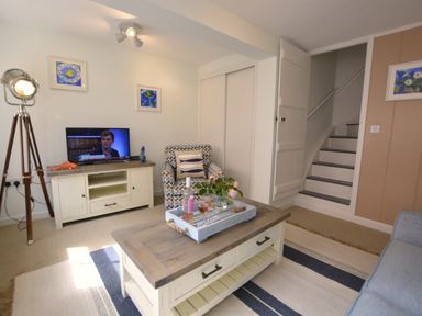 A living room with a coffee table holding a tray with flowers and drinks a patterned armchair a television on a cabinet a floor lamp and stairs at Alinka Aldeburgh in Aldeburgh