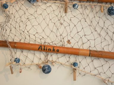 A fishing net with blue floats and shells hanging on a wall with a wooden rod labeled Alinka at Alinka Aldeburgh in Aldeburgh