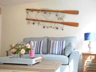 A living room with a blue sofa striped cushions a wooden table with flowers and a bottle on a tray wooden paddles with fishing net on the wall and a lamp on a side table at Alinka Aldeburgh