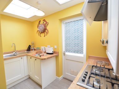 A kitchen with yellow walls a crab wall decoration a countertop with a toaster kettle knives and bread a gas stove and a door with a patterned blind at Alinka Aldeburgh in Aldeburgh