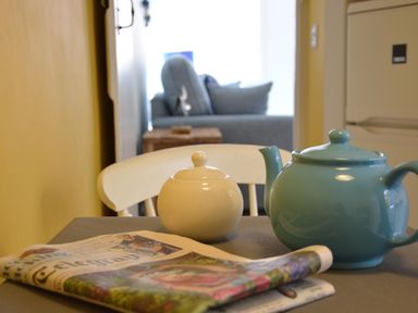 A kitchen table with a blue teapot sugar bowl and a newspaper in a house with a sofa in the background at Alinka Aldeburgh in Aldeburgh