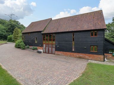 Exterior view of a barn style building with black wooden siding and tiled roof next to a paved driveway and green lawn at Quill Farm Barn in Campsea Ashe