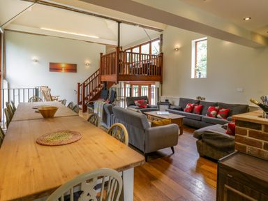 A dining area with a long wooden table and chairs next to a sitting area with grey sofas and red cushions under a wooden staircase at Quill Farm Barn in Campsea Ashe