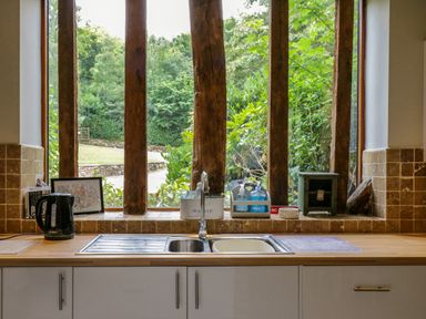 A kitchen sink area with a faucet kettle soap bottles and a window with wooden beams and greenery outside at Quill Farm Barn in Campsea Ashe