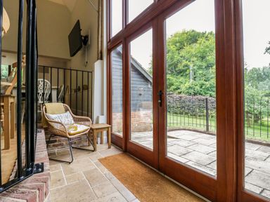 A room with wooden framed glass doors opening to a patio a wicker chair with cushions a small wooden side table and a wall mounted television at Quill Farm Barn in Campsea Ashe