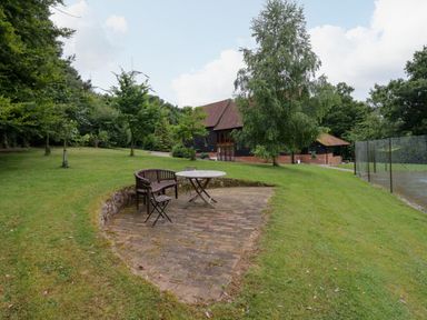 An outdoor seating area with a round table and chairs on a brick patio near a house with trees and a fenced tennis court at Quill Farm Barn in Campsea Ashe