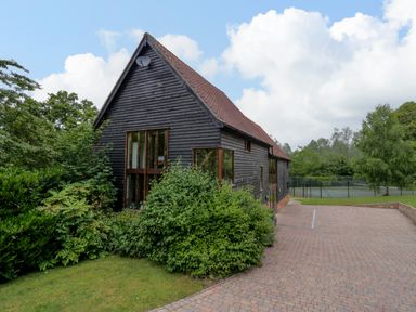 A black wooden barn with large windows surrounded by greenery and a paved driveway with a tennis court in the background at Quill Farm Barn in Campsea Ashe