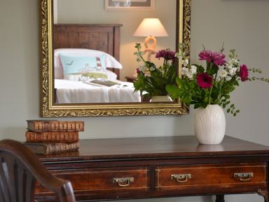 A wooden desk with old books and a vase of flowers in front of a mirror reflecting a bed and lamp at Church Farm Dunwich Near Westleton