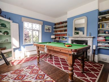 A room with a pool table in the center, bookshelves, a fireplace with a mirror, and a window with diamond-shaped panes at Church Farm Dunwich Near Westleton