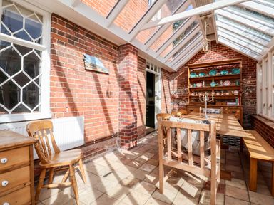 A dining area with wooden chairs a table a bench and a cabinet with dishes in a room with brick walls and a glass ceiling at Church Farm Dunwich Near Westleton