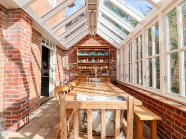 A dining area with a wooden table and chairs under a glass roof with brick walls and a wooden cabinet at Church Farm Dunwich Near Westleton