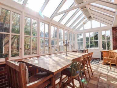 A sunlit dining room with a wooden table and chairs surrounded by glass windows at Church Farm Dunwich Near Westleton