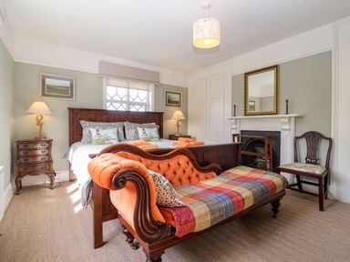 A bedroom with a wooden bed, an orange upholstered bench, bedside tables with lamps, a fireplace with a mirror, and a wooden chair at Church Farm Dunwich Near Westleton