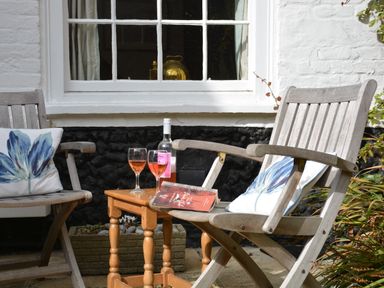 Two wooden chairs with floral cushions and a wooden table with a bottle of wine two glasses and a book outside a white brick building with a window at Mariners Cottage King Street in Aldeburgh
