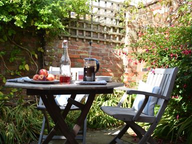 An outdoor wooden table and chairs with a bottle, fruit, and coffee press in a garden at Mariners Cottage King Street Aldeburgh